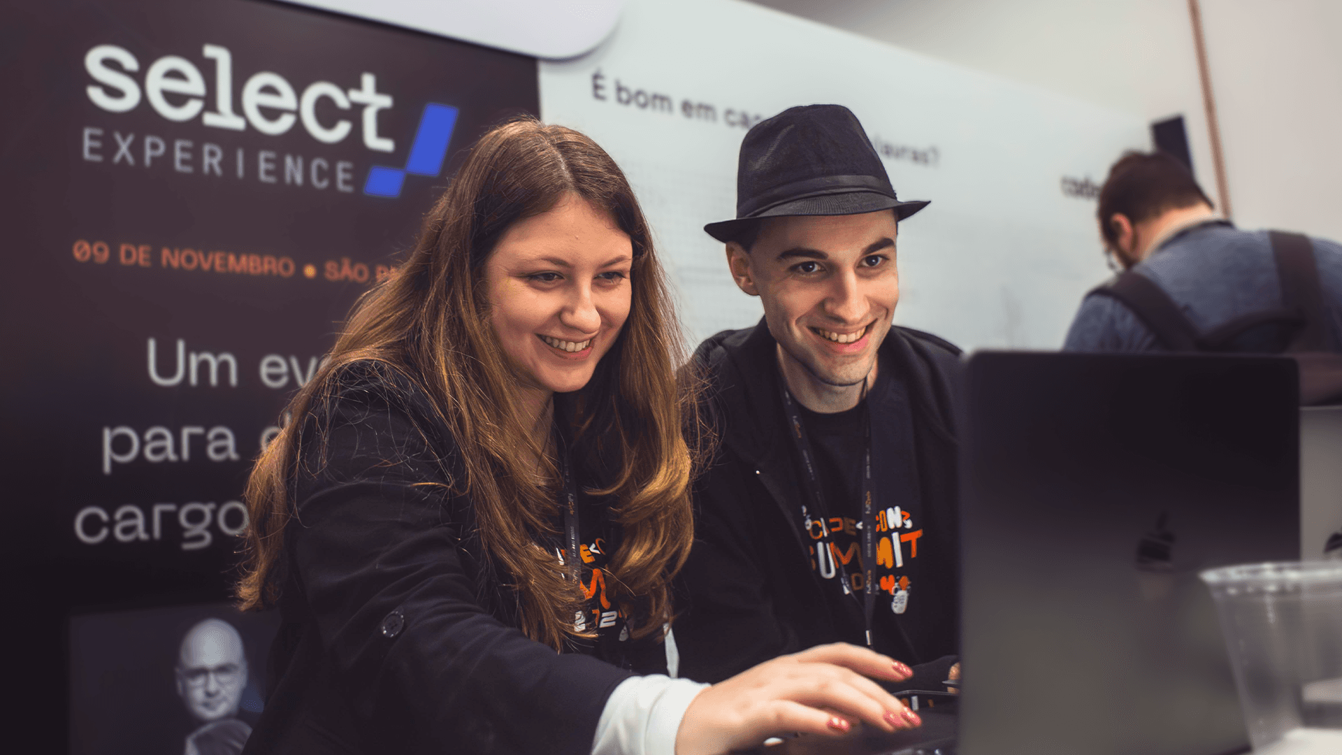 Dois desenvolvedores de software sorrindo enquanto trabalham em um notebook em evento de tecnologia, com banner ao fundo destacando o 'Select Experience', uma conferência voltada para inovações no setor.