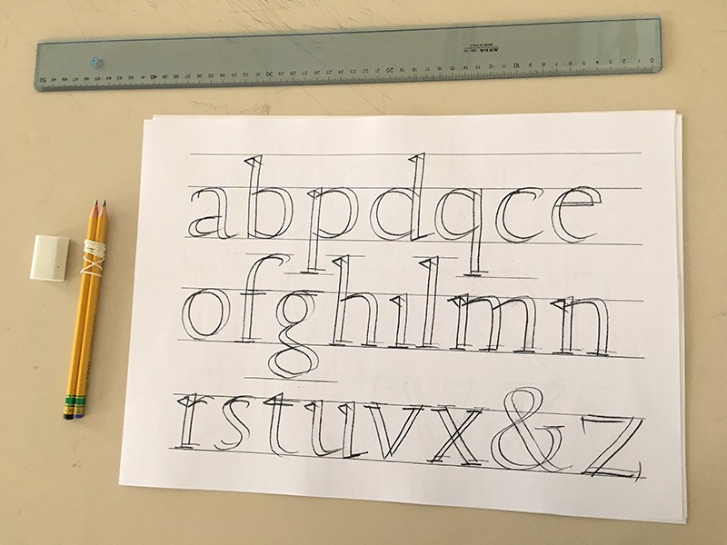 A ruler, two pencils tied, rubber and a paper depicting the alphabet written with the double pencils.