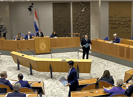 A decorative image showing the plenary hall of the House of Representatives of The Netherlands. The chamber features rows of green seats arranged in a semicircular pattern facing a central podium.