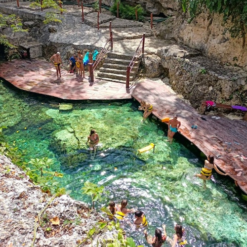 People swimming and relaxing in a crystal-clear natural pool surrounded by rocky cliffs and a wooden platform with stairs.