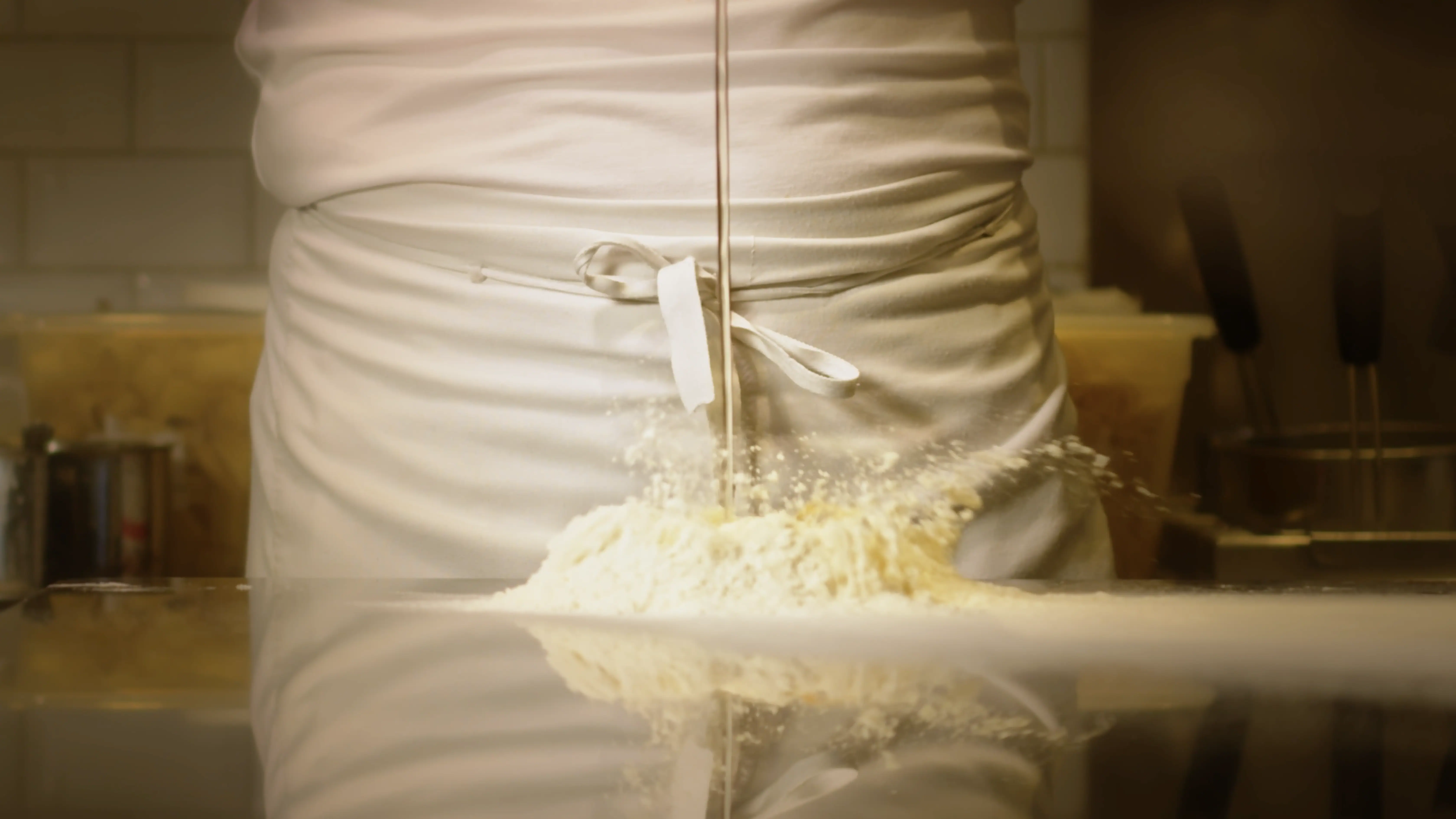 A chef in a white apron energetically stirs a mound of flour on a reflective steel countertop, creating a cloud of flour with cooking utensils blurred in the background.