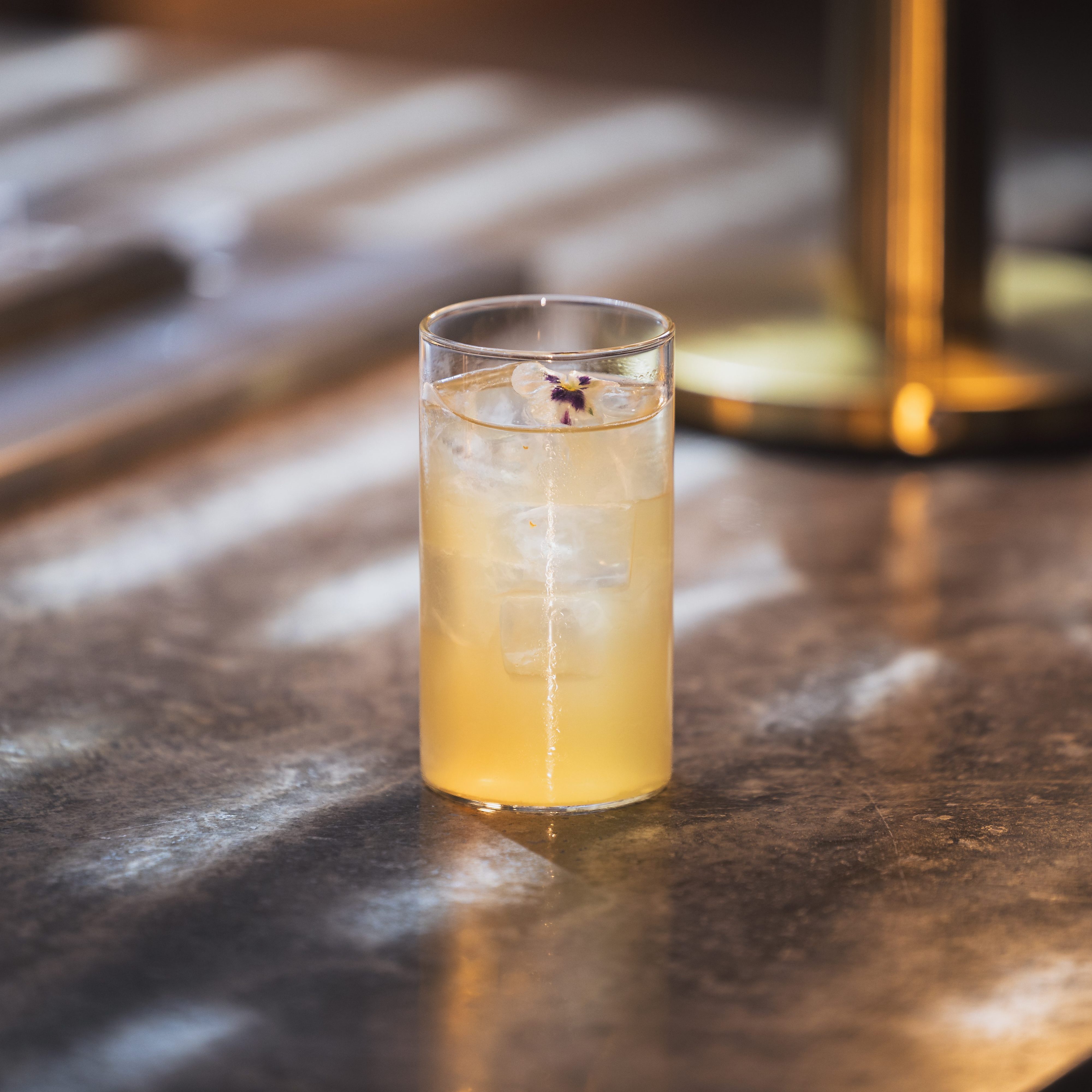 A glass of cocktail on a reflective surface, showcasing a light-colored drink with ice, garnished with a flower, illuminated by warm light.