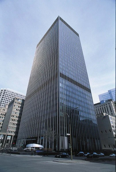 Tall skyscraper with reflective glass windows, set against a clear blue sky. Surrounding buildings frame the scene, conveying a sense of urban modernity.