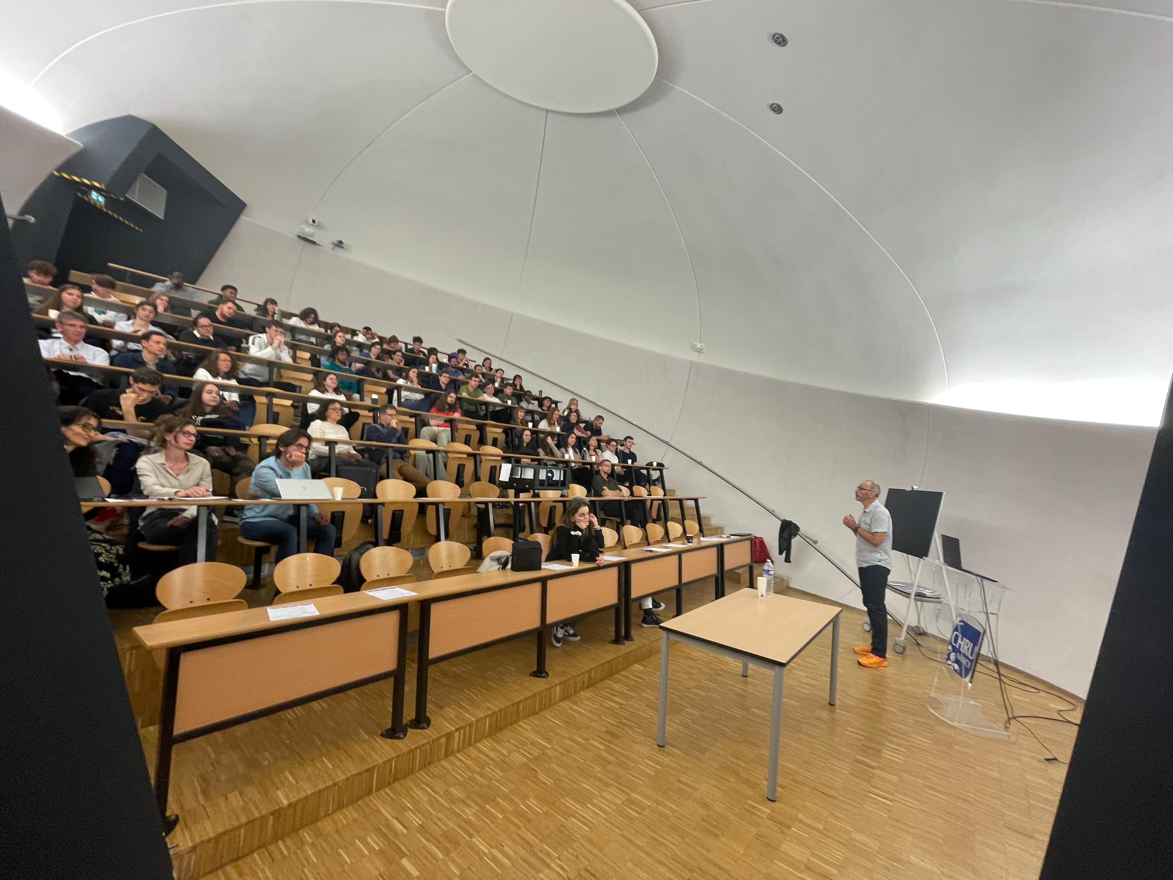Salle de conférence vue en hauteur, occupée par un public assis face à une scène.