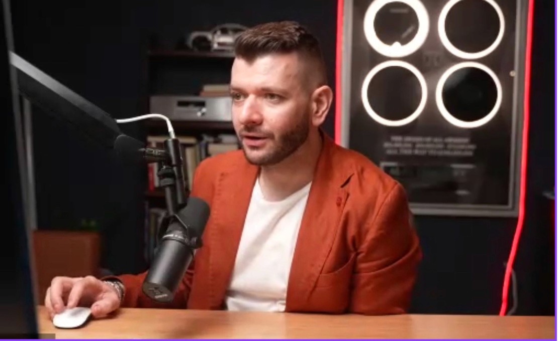Ein Mann im orangefarbenen Sakko spricht in ein Studiomikrofon, ein Standbild aus dem Videointerview über Zahlungen.