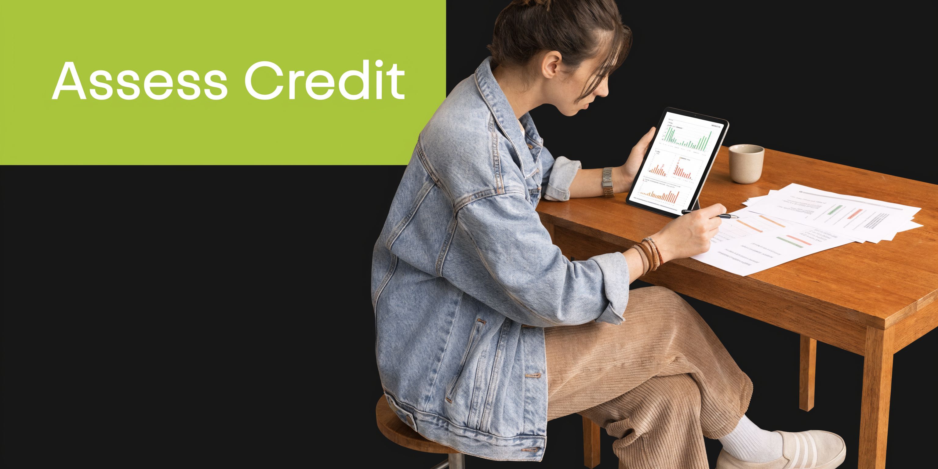 A young woman in a denim jacket analyzes financial graphs on a tablet while sitting at a desk.