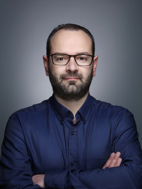 A person with blond hair wears a dark coat and a beige turtleneck sweater. They have a calm expression, against a neutral background.