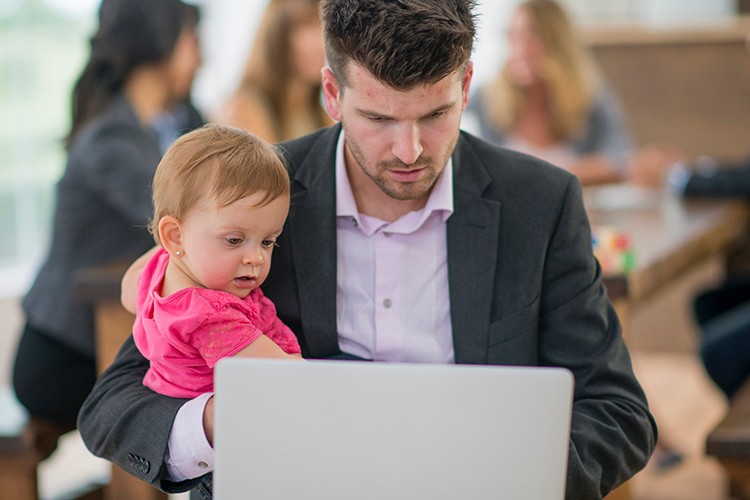 A man in business attire holds a young toddler wearing a bright pink hoodie while working on a laptop in an office setting. Colleagues are visible but blurred in the background, illustrating work-life balance and a family-friendly workplace environment that accommodates working parents.