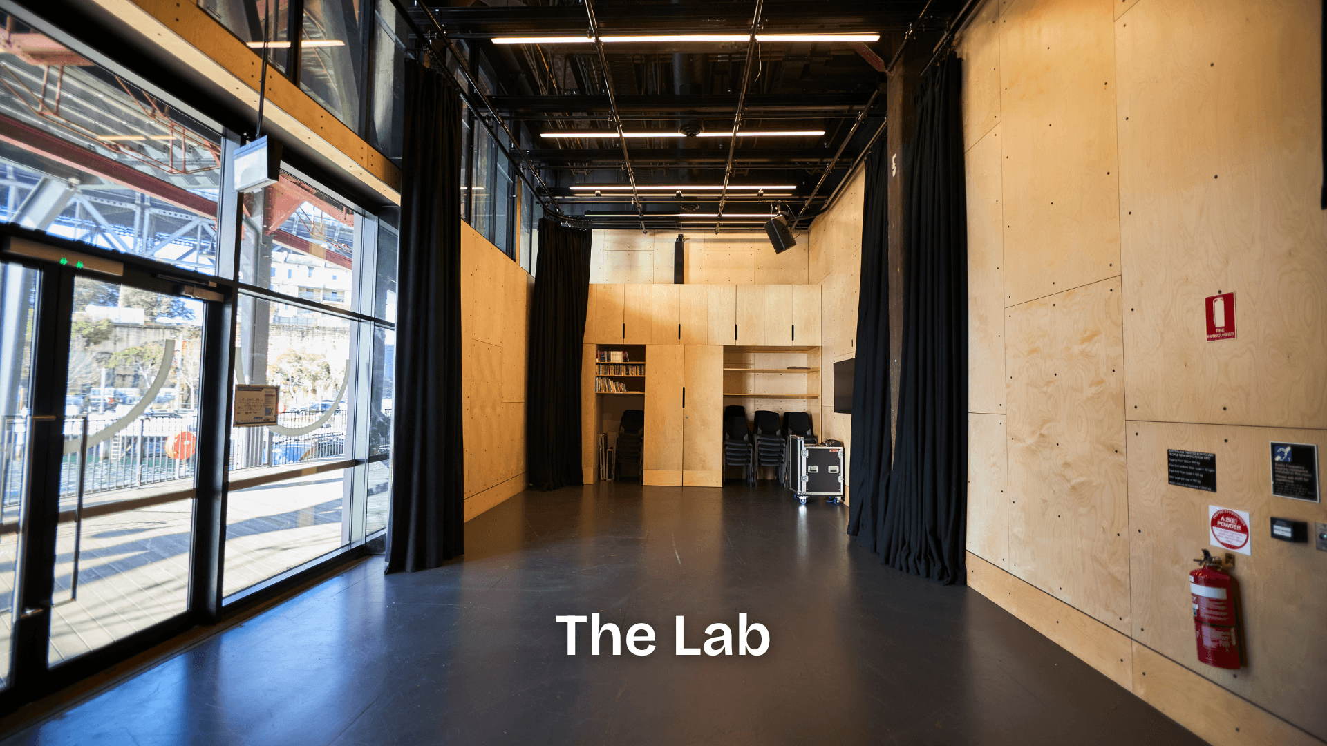 A light-filled rehearsal studio with timber walls, tall windows, black curtains, and stacked chairs, set up for drama workshops and creative development.