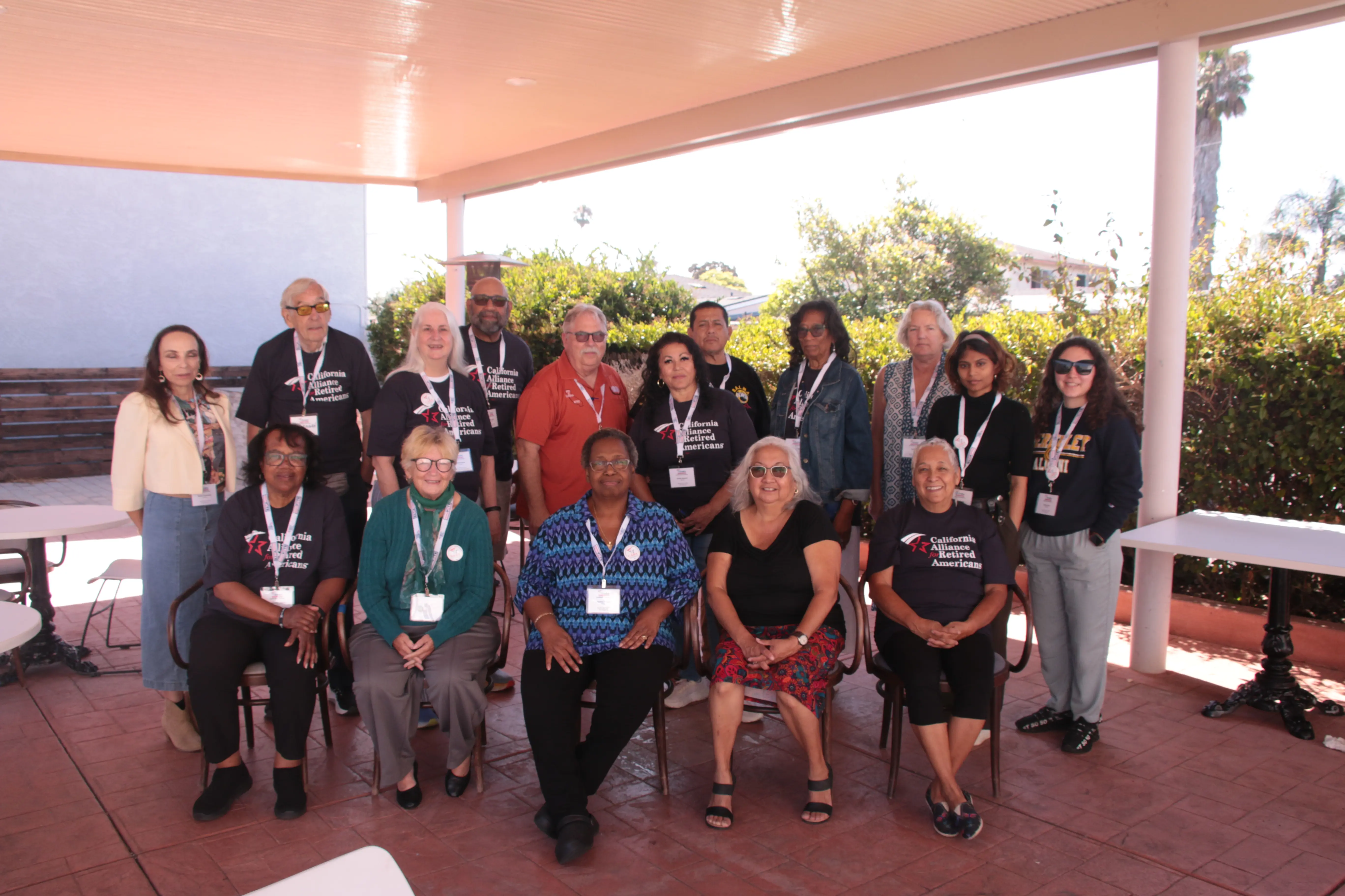 A diverse group of people, many wearing “California Alliance for Retired Americans” shirts and lanyards, pose together for a group photo under a covered outdoor area.