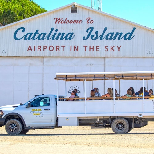 A tour vehicle with passengers is parked in front of a building with the sign, "Welcome to Catalina Island Airport in the Sky."