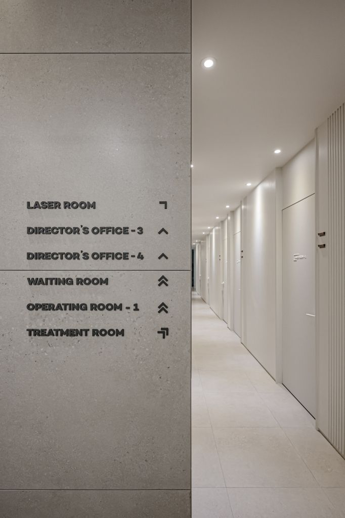Minimalist medical clinic wayfinding sign with black directional lettering listing laser room, director’s office, waiting room, operating room, and treatment room along a clean hallway.