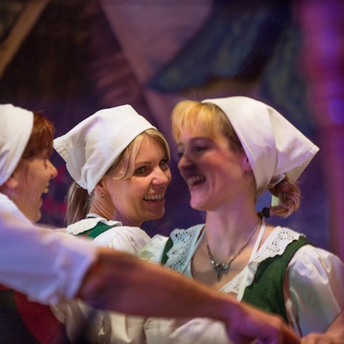 Três mulheres usando lenços brancos e roupas tradicionais sorriem alegremente enquanto dançam juntas.