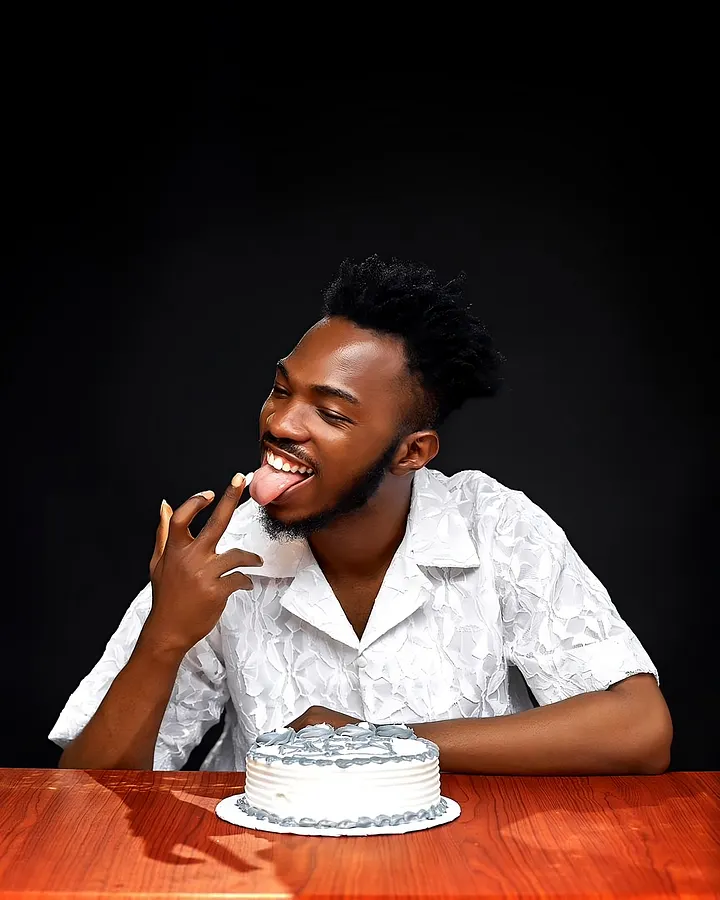 Aremu sits at a table in a white floral shirt and licks his finger next to a cake