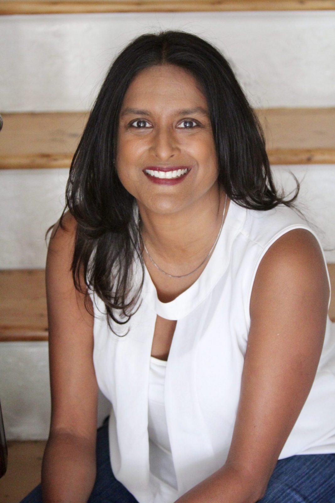 A smiling woman with long dark hair is sitting in front of a wooden backdrop, wearing a white top and a necklace.