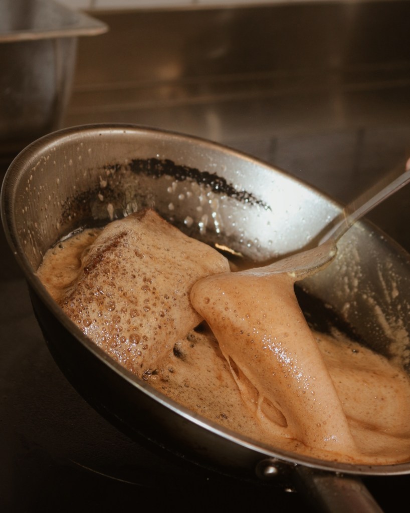 A close-up of a pan with bubbling, frothy liquid, indicating a cooking process, possibly featuring a dark sauce or broth.
