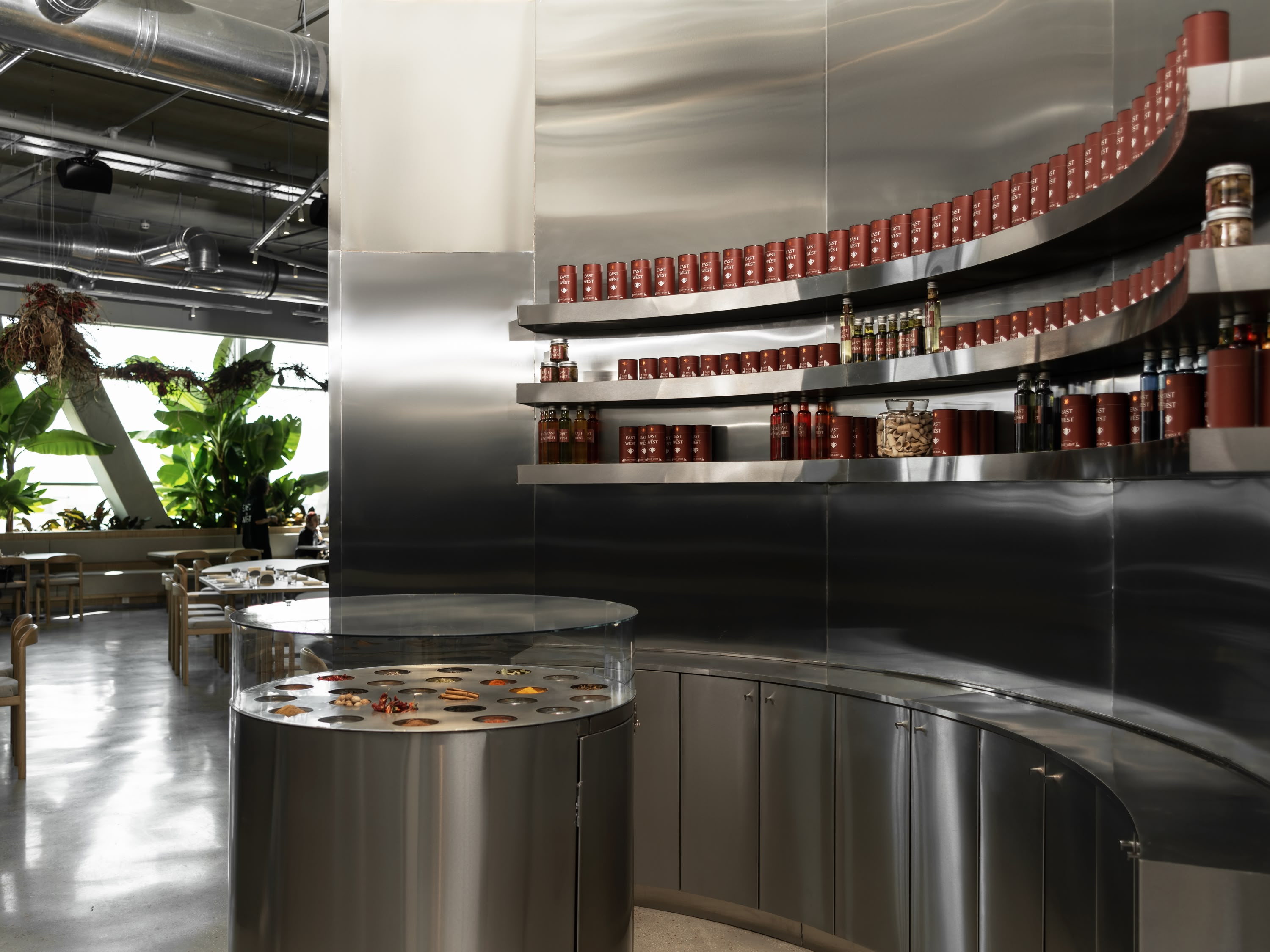 East by West restaurant bar area with curved stainless steel shelving stocked with red jars and bottles, potted plants, hanging vines, round display table amid industrial ducts.