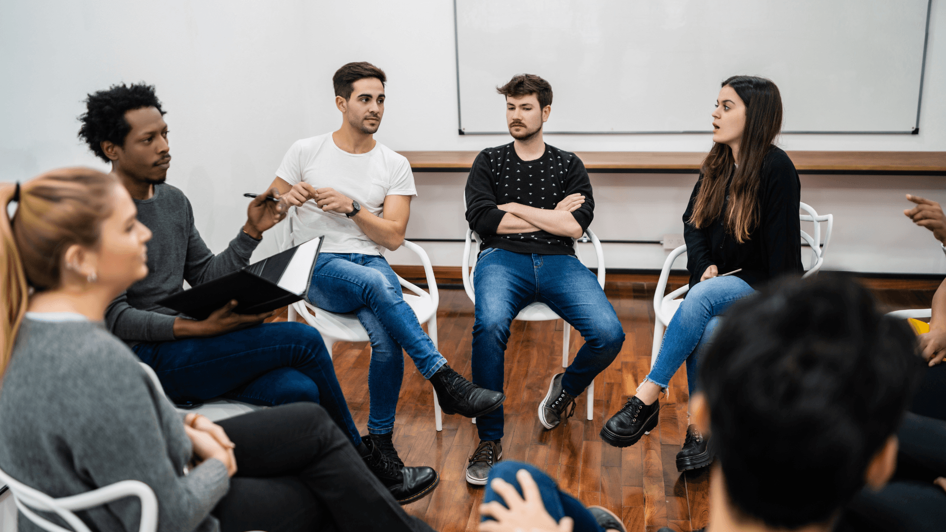 A imagem mostra um grupo de pessoas participando de uma discussão em círculo. Eles estão sentados em cadeiras brancas em uma sala com piso de madeira e um quadro branco ao fundo. O ambiente parece ser uma reunião ou sessão de grupo, com uma atmosfera descontraída, onde cada pessoa participa de forma engajada. Algumas pessoas estão falando, enquanto outras escutam atentamente. A configuração da sala e o estilo de interação sugerem um espaço colaborativo, onde o foco está na troca de ideias ou em uma sessão de apoio mútuo.