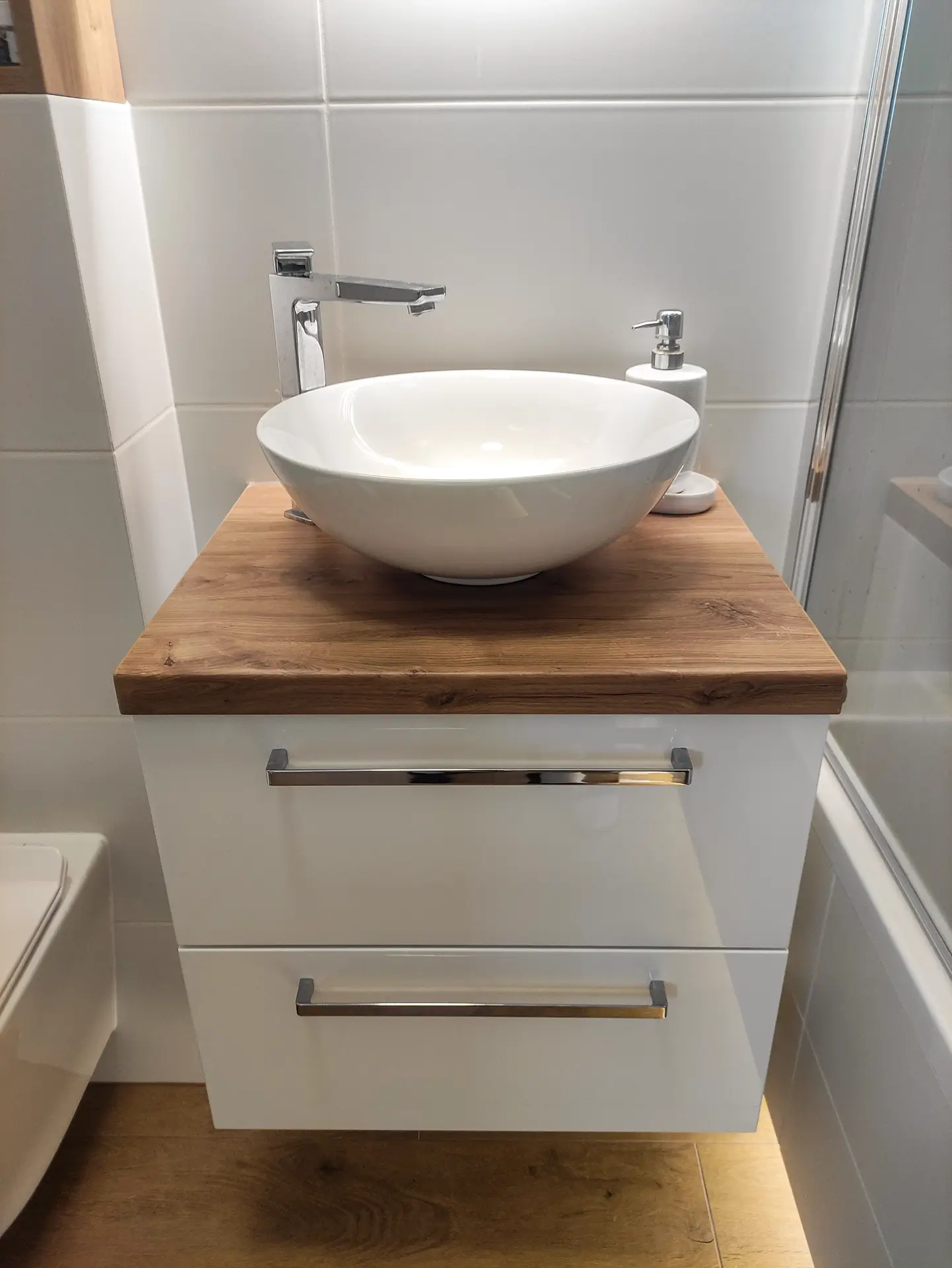 Mała szafka łazienkowa z szufladami, drewnianym blatem i białą umywalką nablatową.