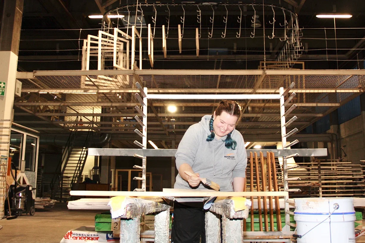 Junge Frau bürstet Holz in Werkstatt, gestelltauf Holzwerkstück, im Hintergrund Gestelle mit Holzteilen.