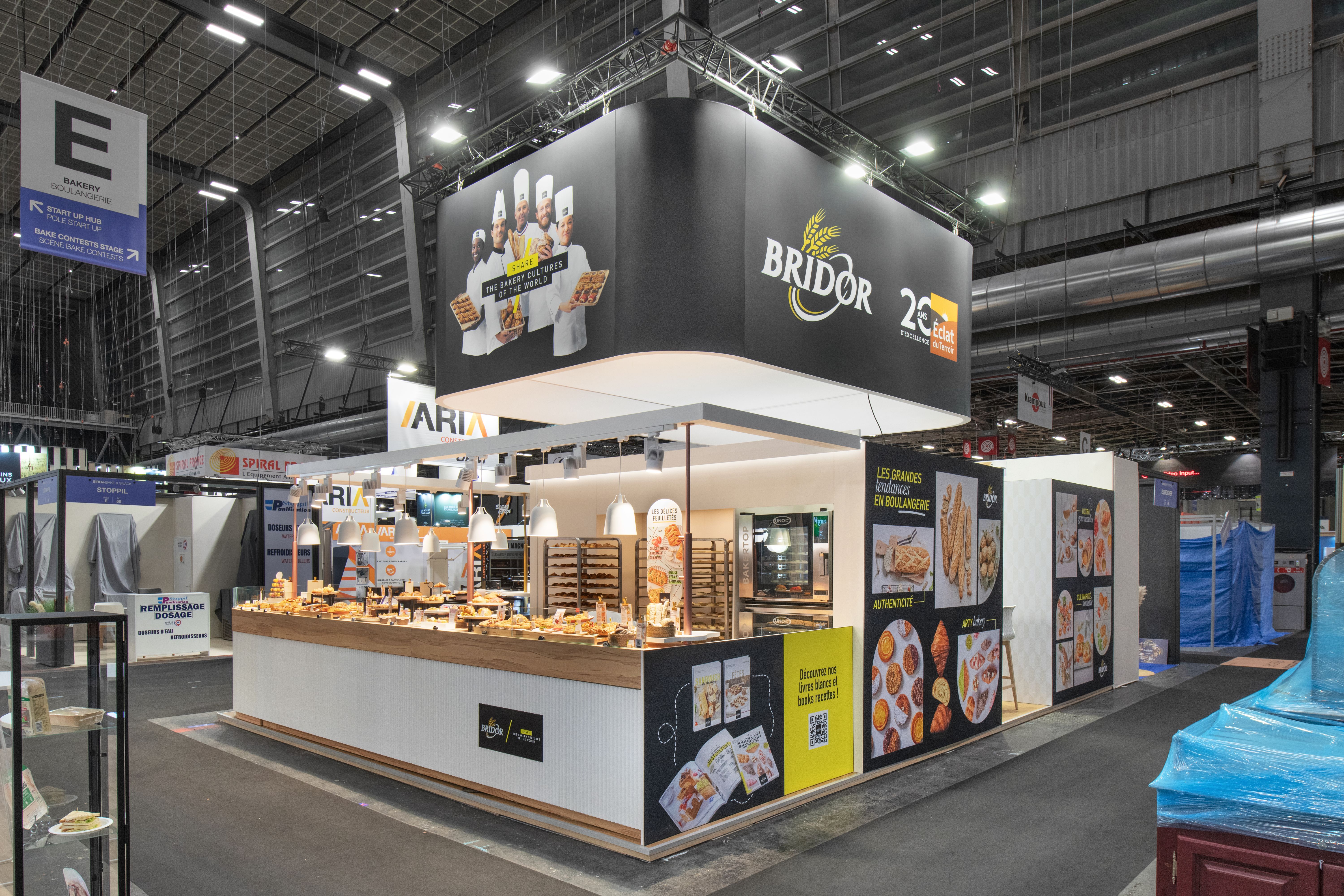 Stand Bridor Bake and Snack 2026 conçu par Expace – vue d'ensemble avec caisson suspendu Share the Bakery Cultures of the World, boulangers en démonstration au comptoir et mur de visuels viennoiseries