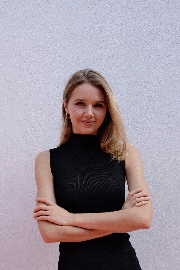 Portrait of me in a sleeveless black top, standing with arms crossed and a slight smile, in front of a textured pale wall.