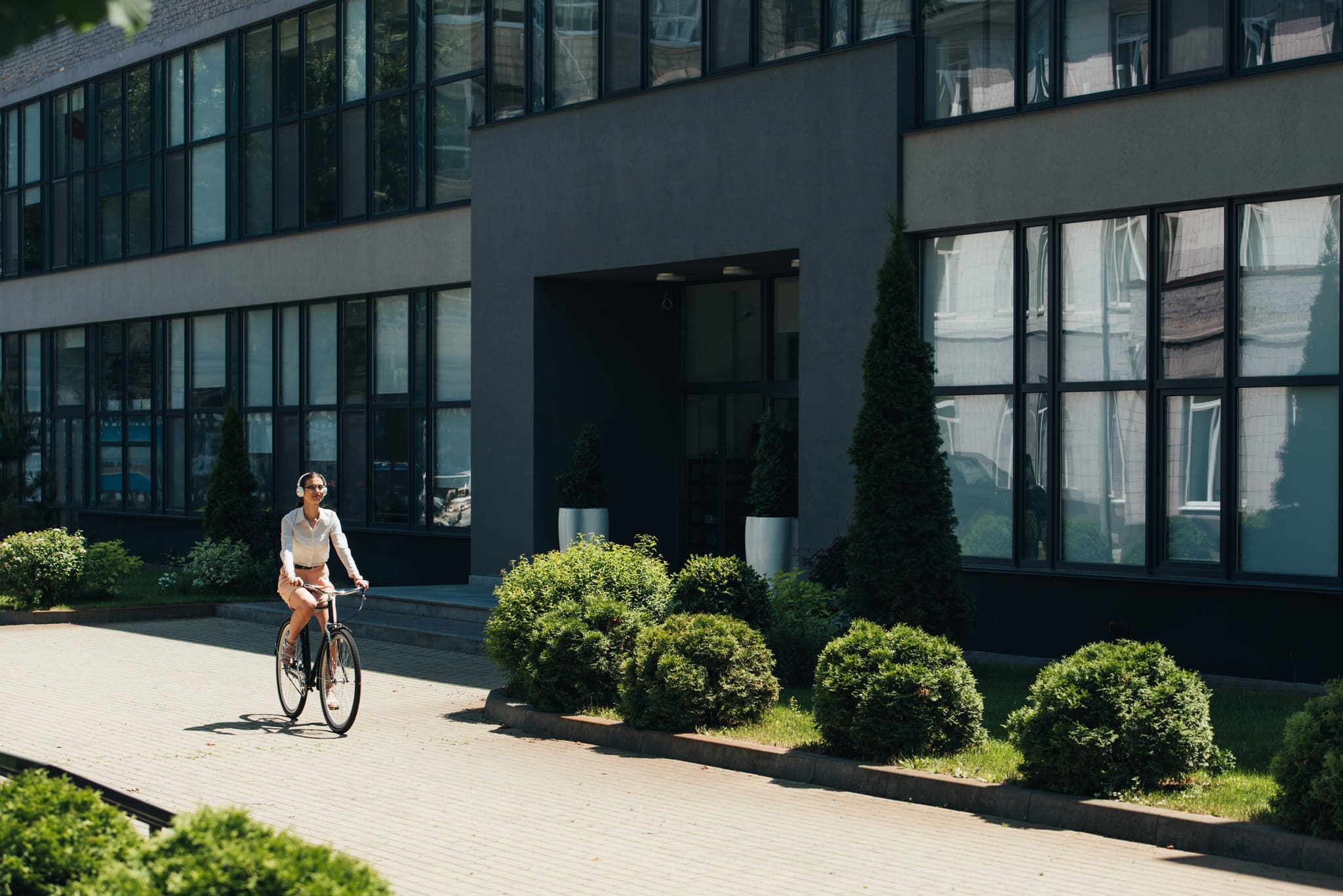 A person wearing headphones riding a bicycle on a sunny path in front of a modern, dark-facade office building with green hedges.