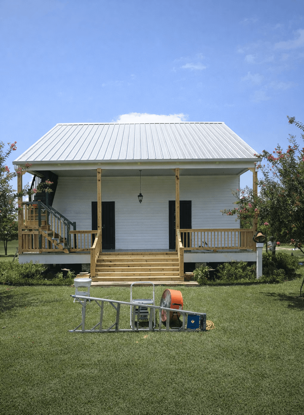 Two-story white house with a wraparound balcony overlooks a clear blue swimming pool. Palm trees and potted plants add a tropical vibe under a sunny sky.