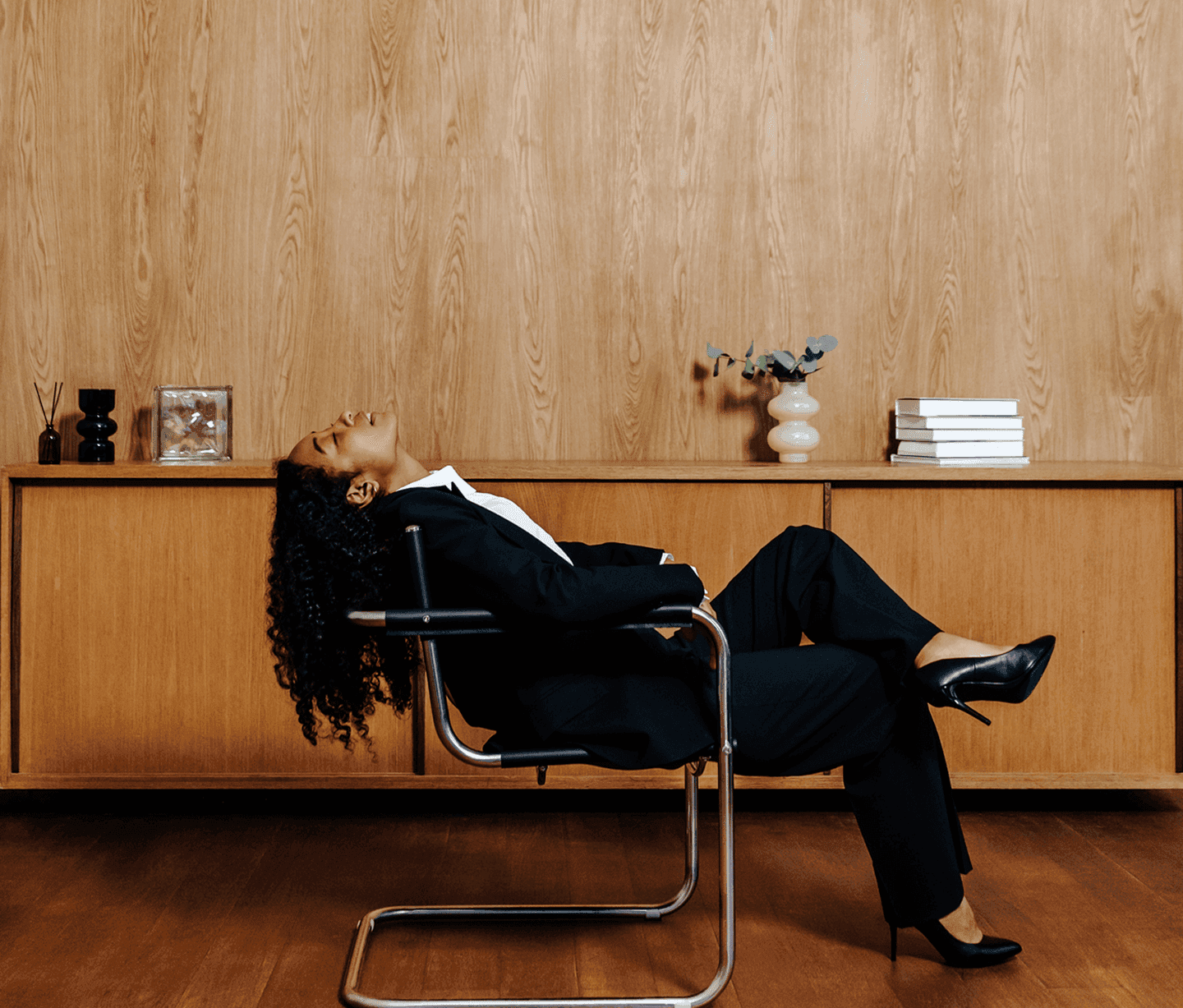 Businesswoman in a sleek black suit reclining in a modern chrome chair, set against a wooden cabinet with minimalist decor, exuding a relaxed and confident vibe.