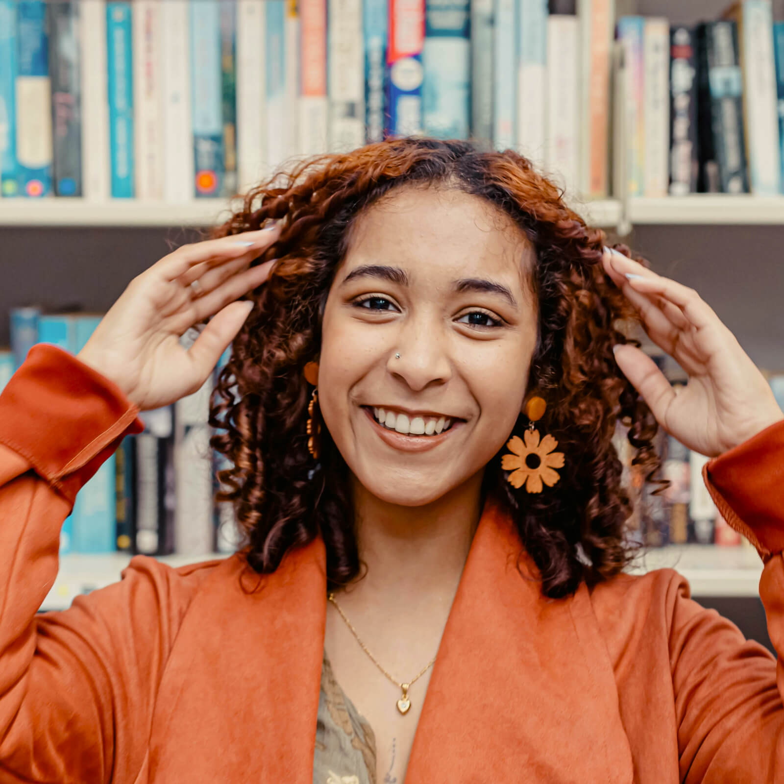 Eine junge Frau mit orangenem Oberteil schaut leicht lächelnd während sie die Hände zum Kopf führt