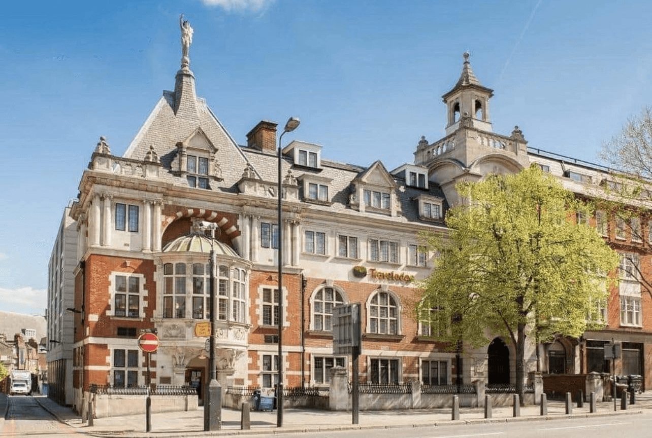 Smiling young woman with long hair standing against a dark green background, holding a finger to her chin.