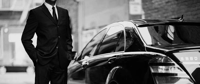 A professional chauffeur in a black suit and white gloves opening the door of a luxury black car for a passenger.