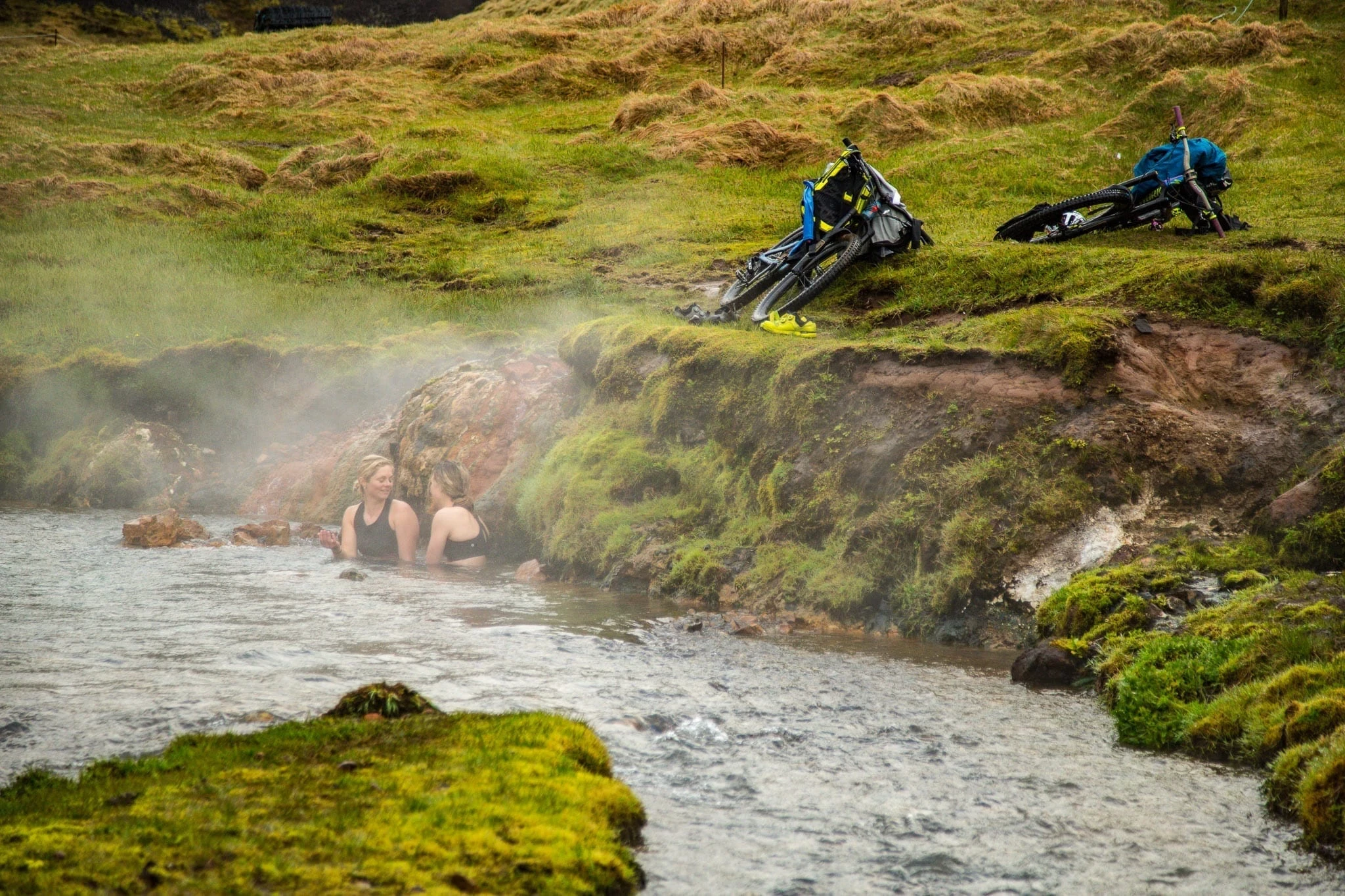 People relaxing in a steaming hot spring, with bikes resting on the bank.