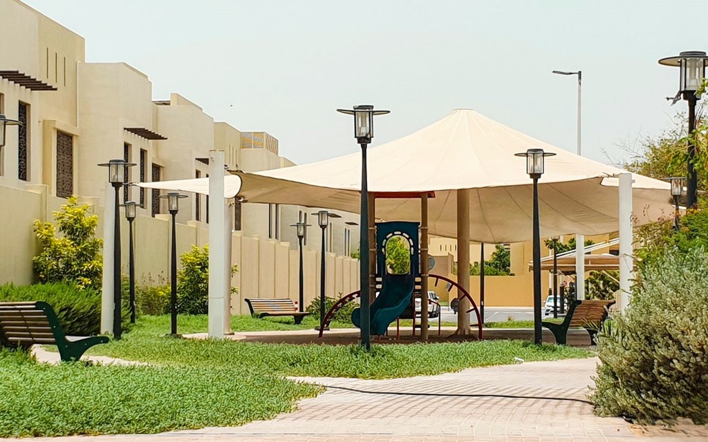 A park in Al Forsan Village featuring a playground and a tent, surrounded by greenery and open space.