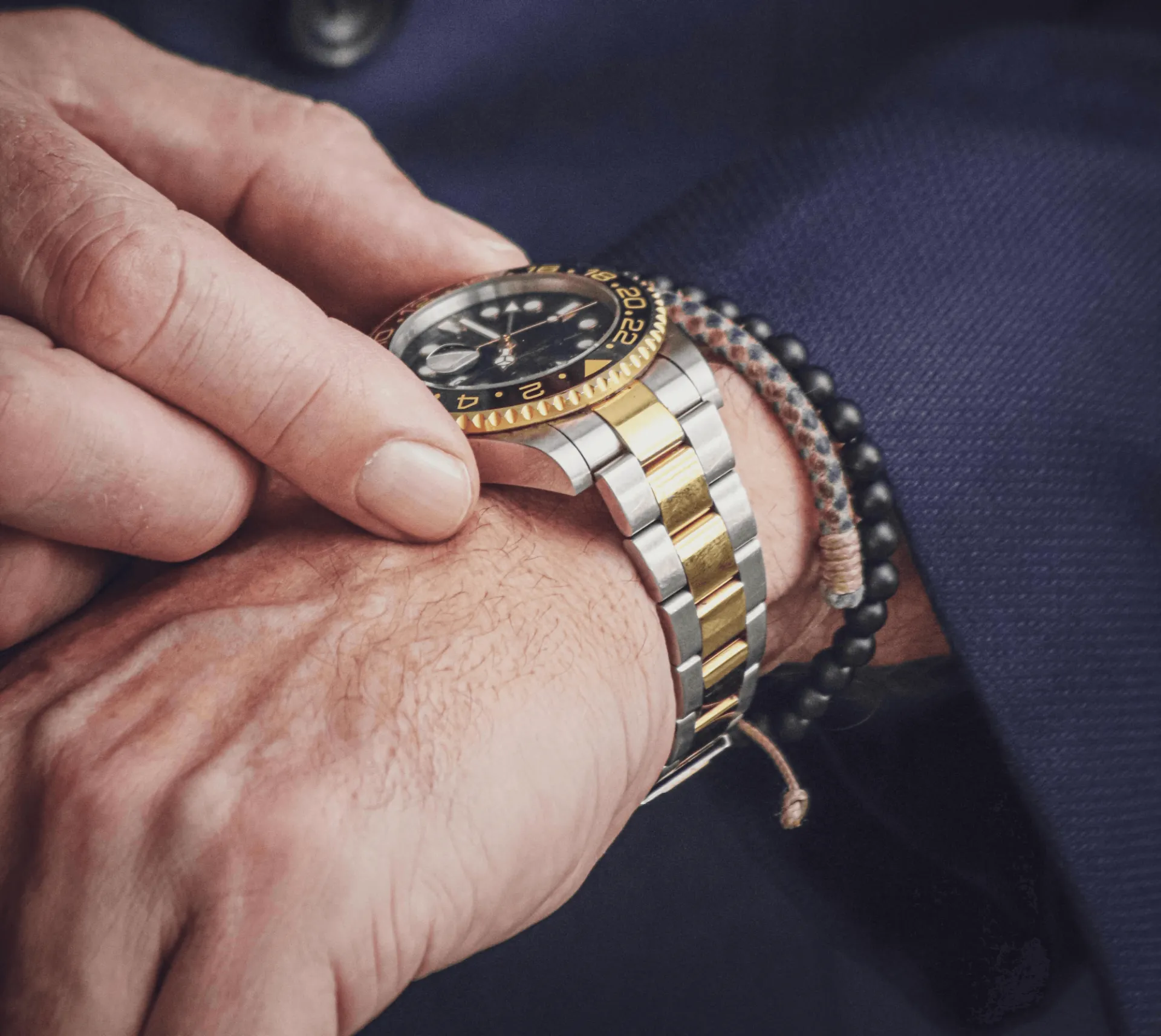 Two-tone Rolex Submariner watch with black dial, gold rotating bezel, and two-tone Oyster bracelet, worn on a wrist alongside a black beaded bracelet against a navy blue suit.