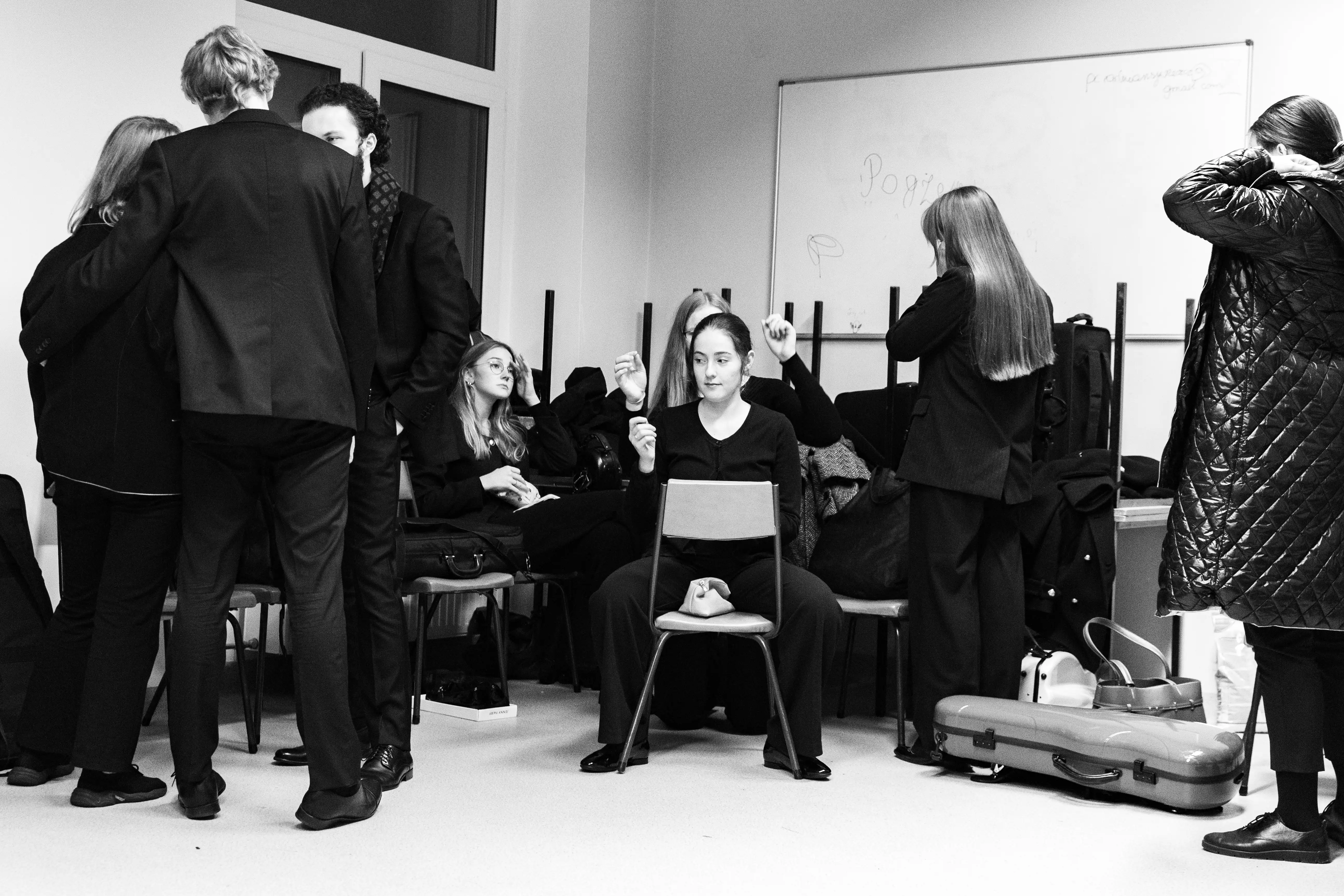 Grupa osób w czarnych strojach w pomieszczeniu, niektórzy rozmawiają, inni przygotowują się do występu. W centrum siedzi kobieta na krześle z zamkniętymi oczami.