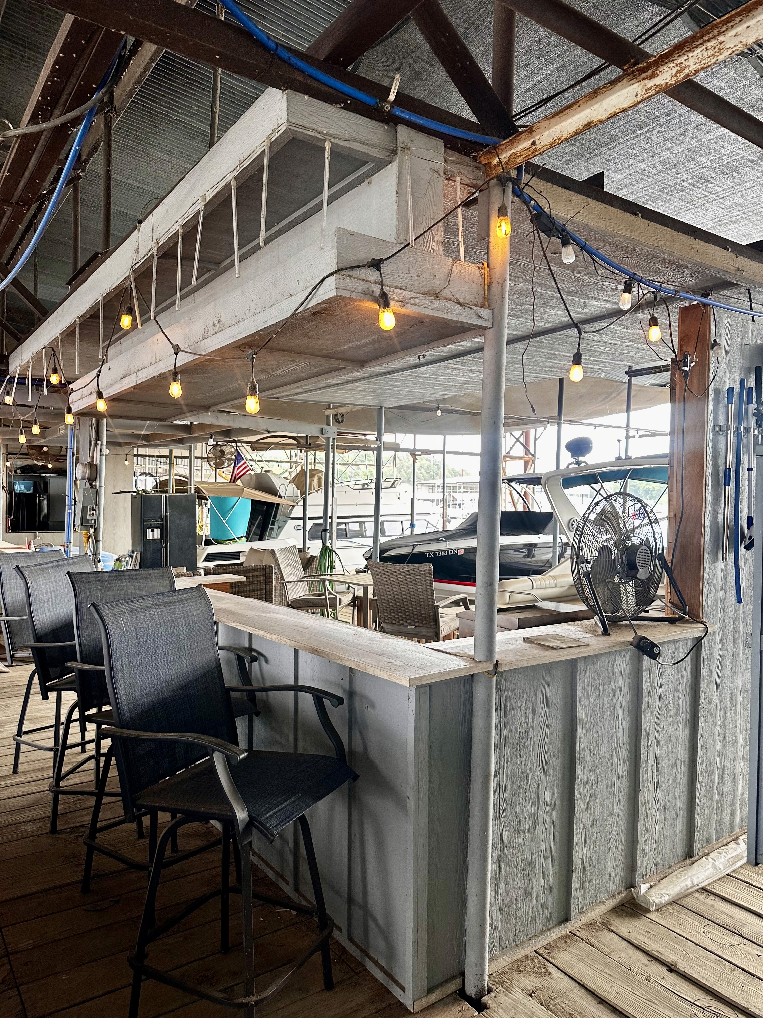 A rustic dockside bar features high chairs, a fan, and hanging string lights, creating a cozy waterfront atmosphere with moored boats visible outside.