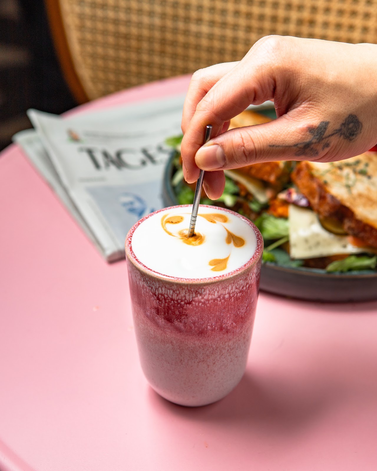 Nahaufnahme einer rosa Keramik-Tasse mit Latte Art, die mit einem Metalllöffel umgerührt wird, mit einem unscharfen Sandwich und Salat im Hintergrund.