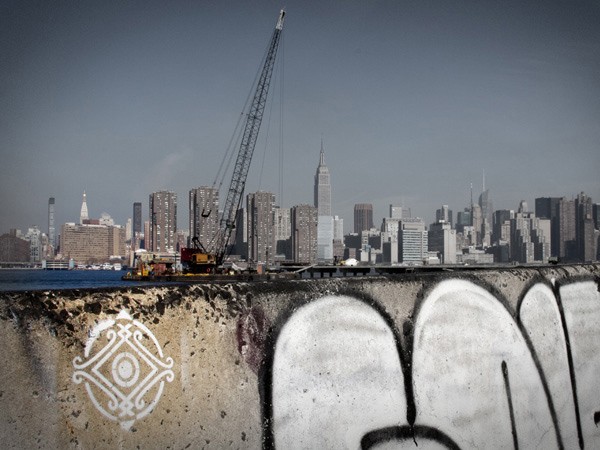 A construction crane is positioned on a platform overlooking the New York City skyline. The crane is dark gray or black and extends upward. The foreground features a concrete wall with graffiti, including a white design on the left side and large, light gray or white letters on the right side. The wall shows signs of wear and discoloration. The background includes the skyline of tall buildings under a clear, light gray or blue sky.