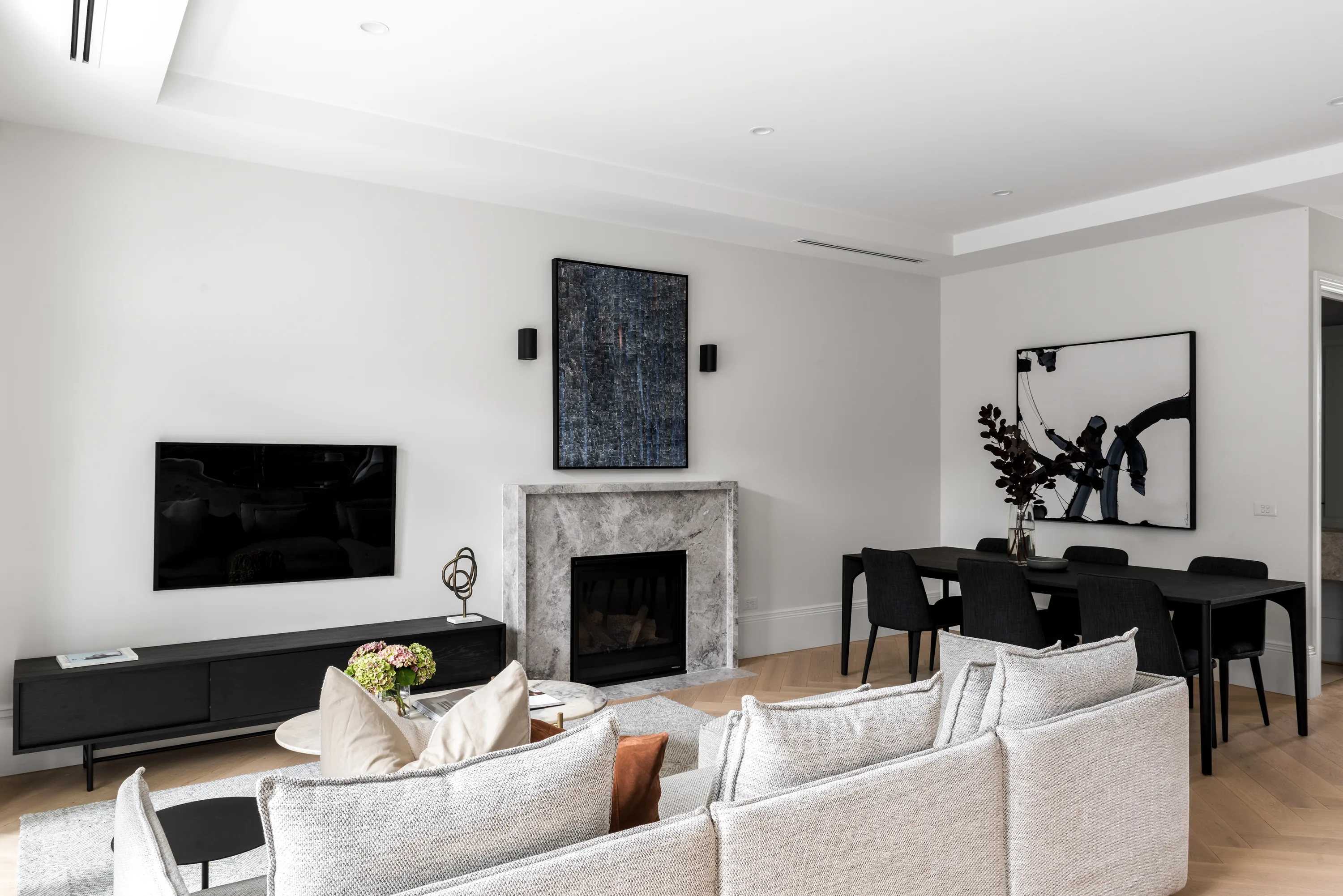 Living and dining area with a grey-marble fireplace, gallery-style artwork, a black media unit and TV, pale sofas and a black dining table with charcoal chairs