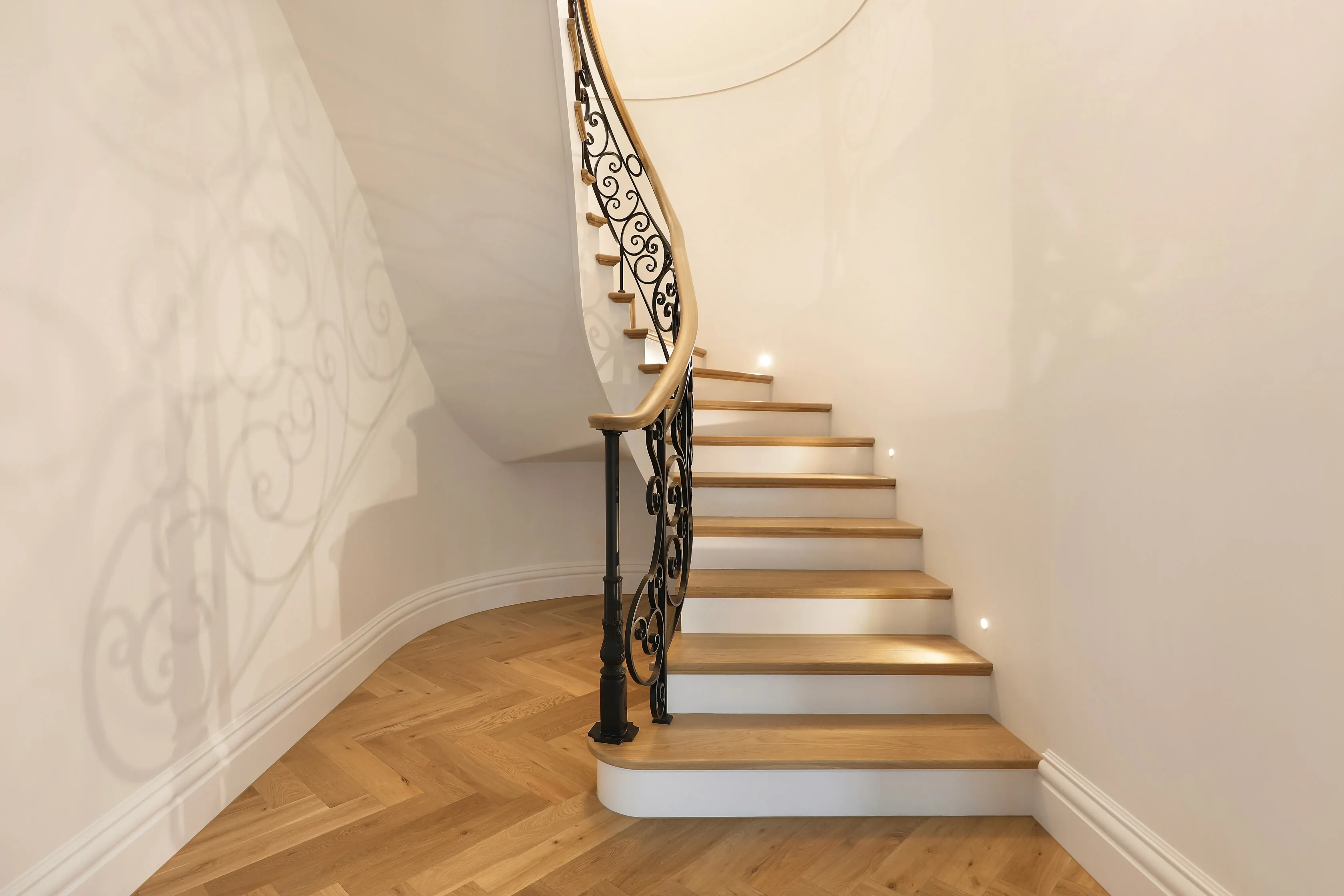 Sculptural curved staircase wrapped in hand-forged black iron lacework with an oak handrail and oak treads, rising over a herringbone timber floor against white walls