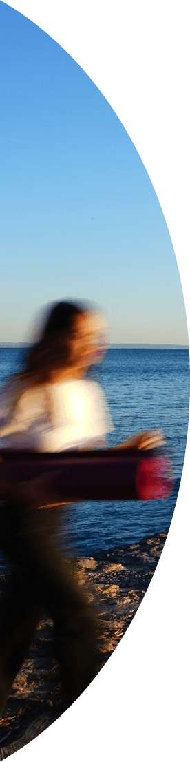 A woman running at the beach with a rolled pilates mat on her right hand.