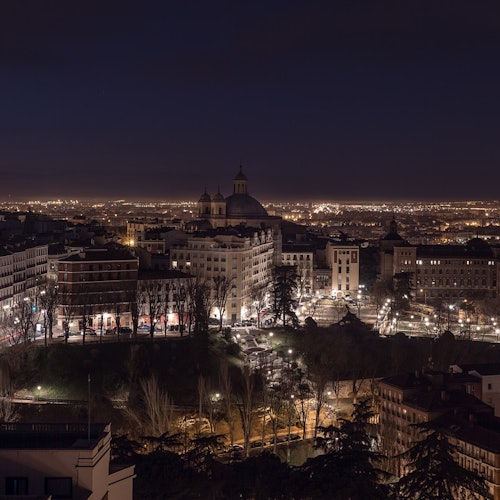 Αστικό τοπίο τη νύχτα με φωτισμένα κτίρια, μια κεντρική περιοχή πάρκου και ένα εξέχον θολωτό κτίριο στο βάθος.
