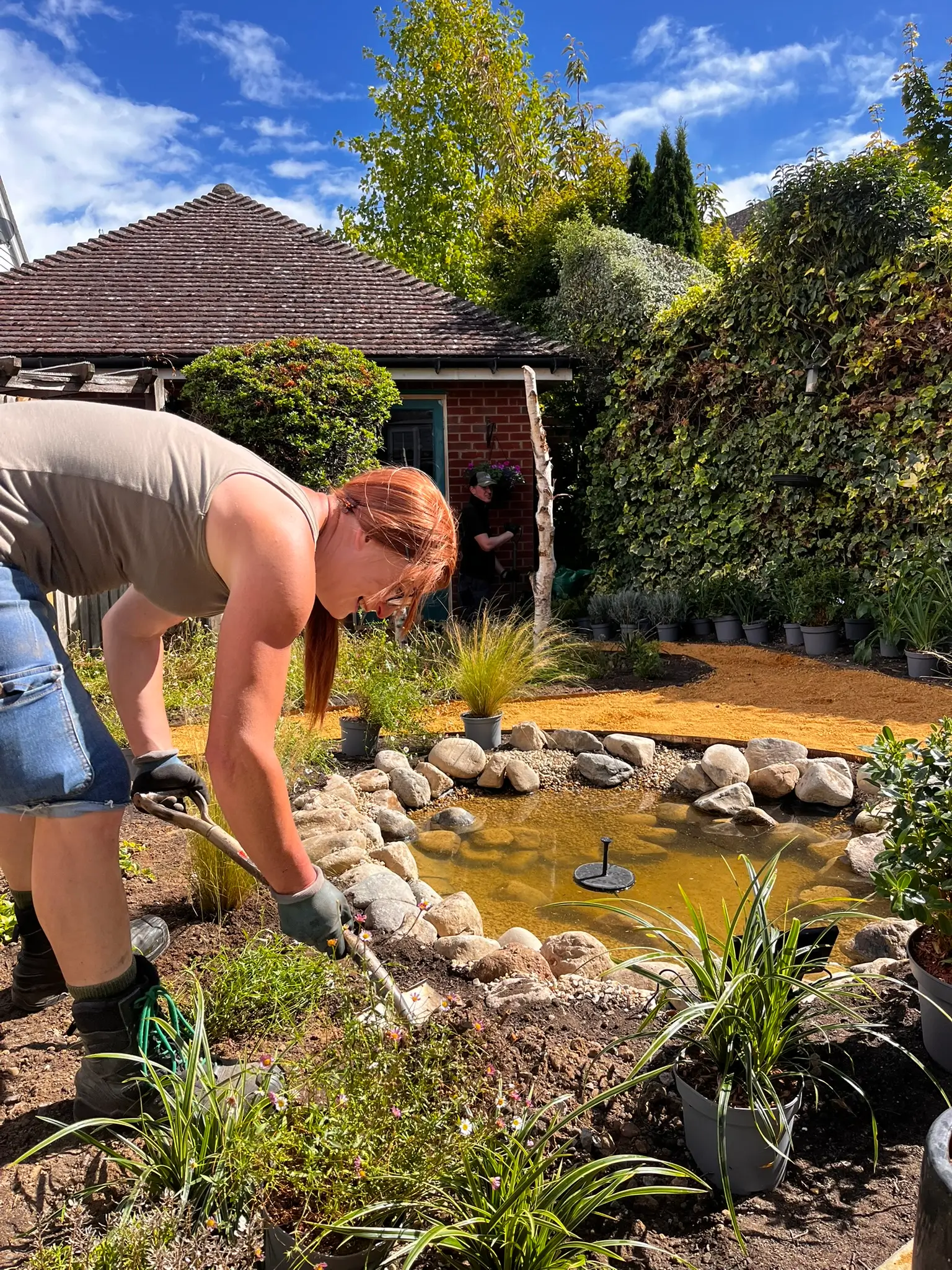 Person tending to a garden pond, surrounded by greenery and blue skies.