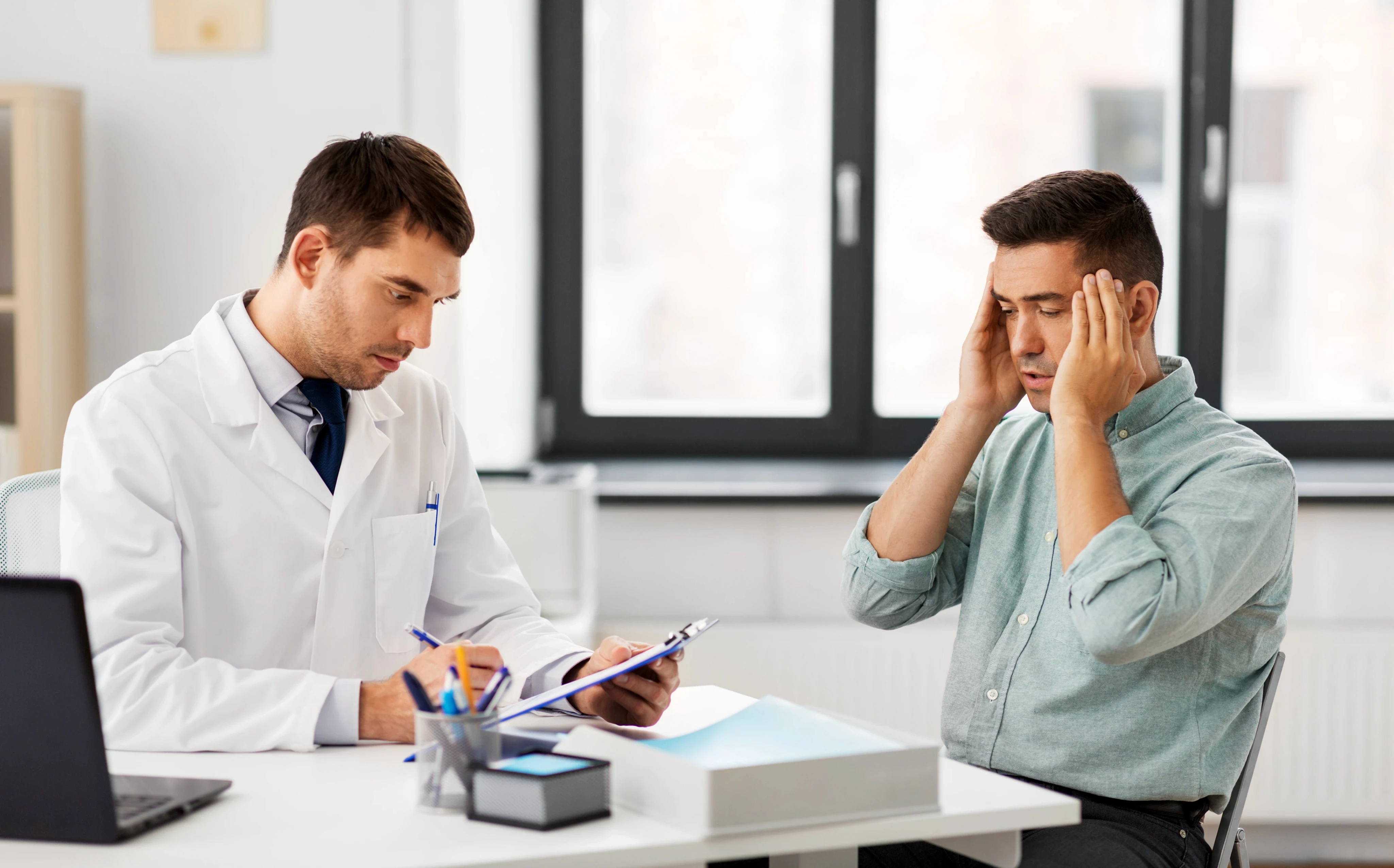 A doctor in a white coat listens attentively to a patient sitting across the desk, who holds his head, appearing stressed or in pain, in a clinical setting.