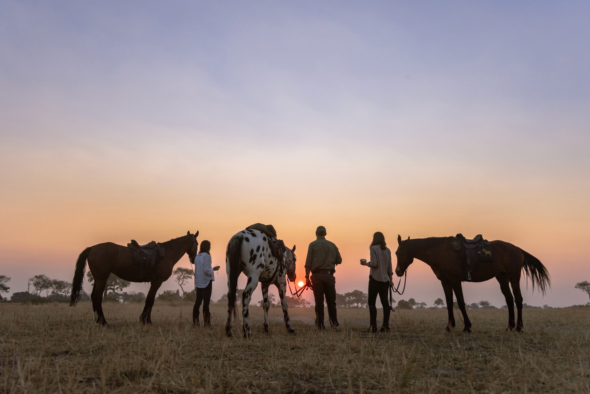 Kujwana Delta Ride, Okavango Delta, Botswana. Tre ryttare står bredvid sina hästar. De är på ridsafari och med en drink i handen blickar de ut över solnedgången.