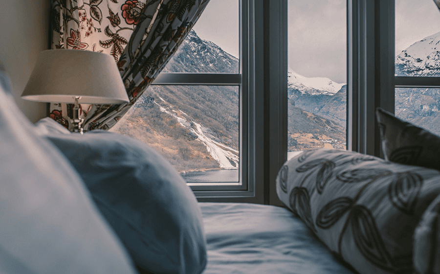 A cozy bedroom with plush pillows and a floral curtain, offering a stunning view of snow-capped mountains through large glass windows, creates a serene and picturesque atmosphere.