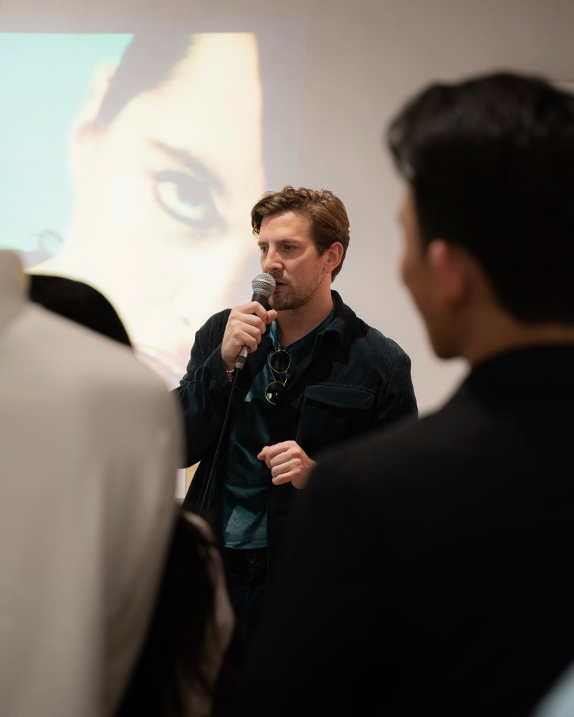 A man speaking into a microphone, wearing a blue jacket and holding sunglasses, with an image of a woman's eye projected in the background. People are partially visible in the foreground.