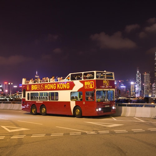 夜晚的城市天际线上,一辆标有“BIG BUS HONG KONG”字样的红白双层巴士,乘客坐在上层。
