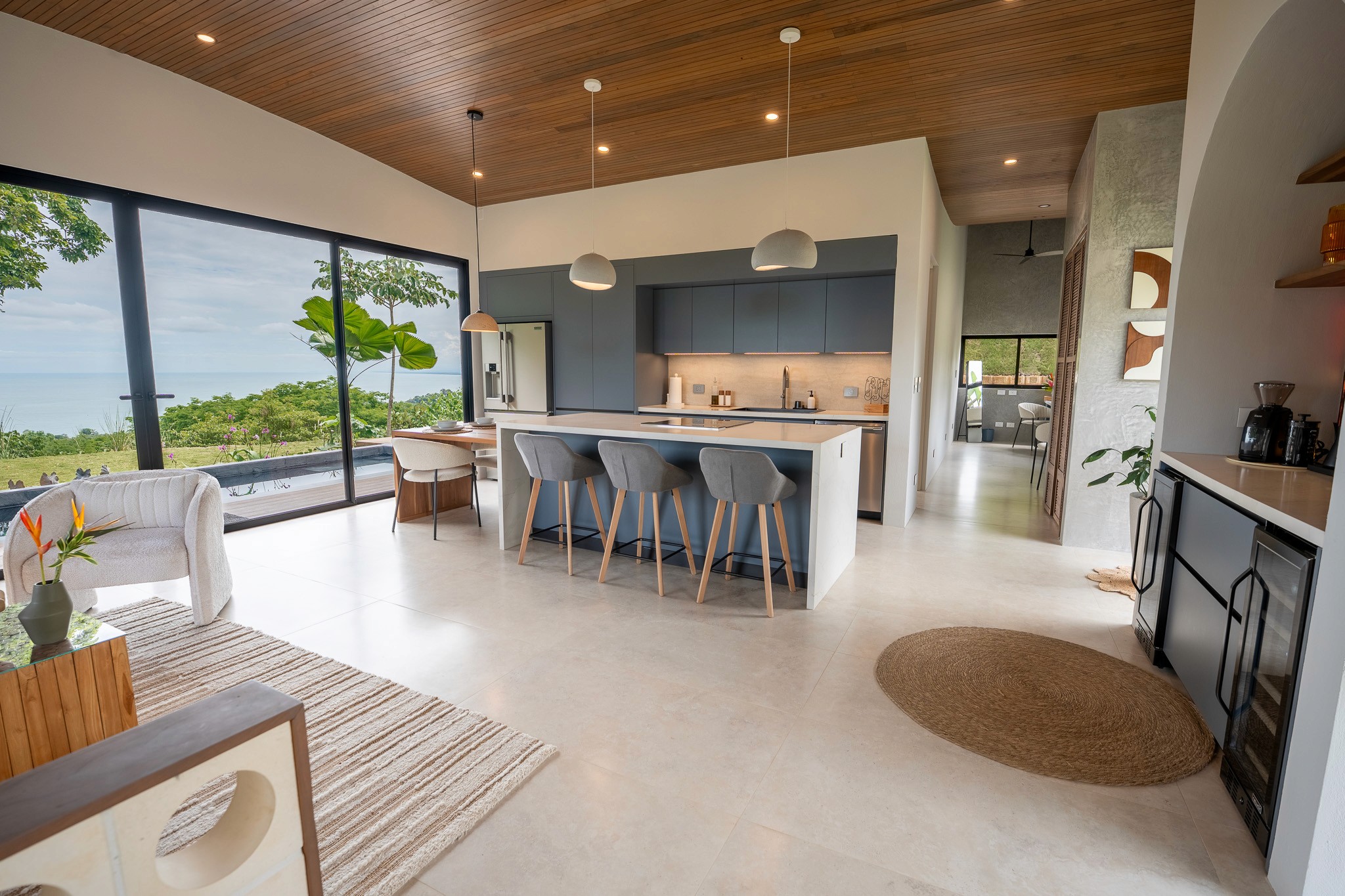 Spacious open-concept living room and gourmet kitchen with soaring wood ceilings and floor-to-ceiling glass walls showcasing panoramic ocean views.